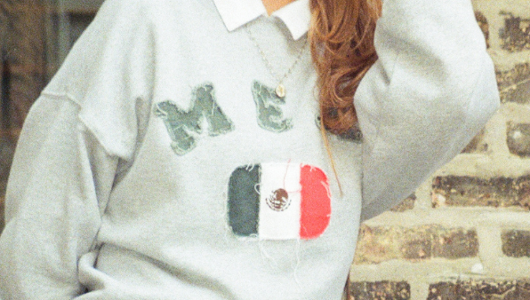 A woman wearing a light gray sweatshirt with a white collar and a Mexican flag graphic on the front, paired with blue jeans and sunglasses, sitting against a brick wall.