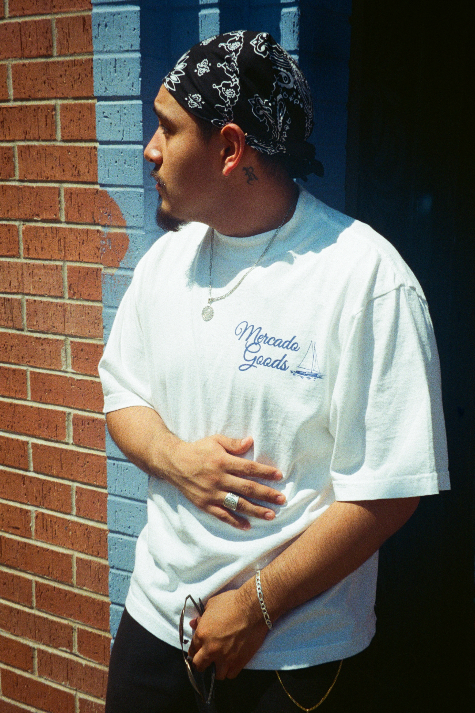 A man wearing a white t-shirt with blue cursive embroidery that reads 'Miranda Goedde', paired with a black and white bandana, standing against a brick and blue wall background.