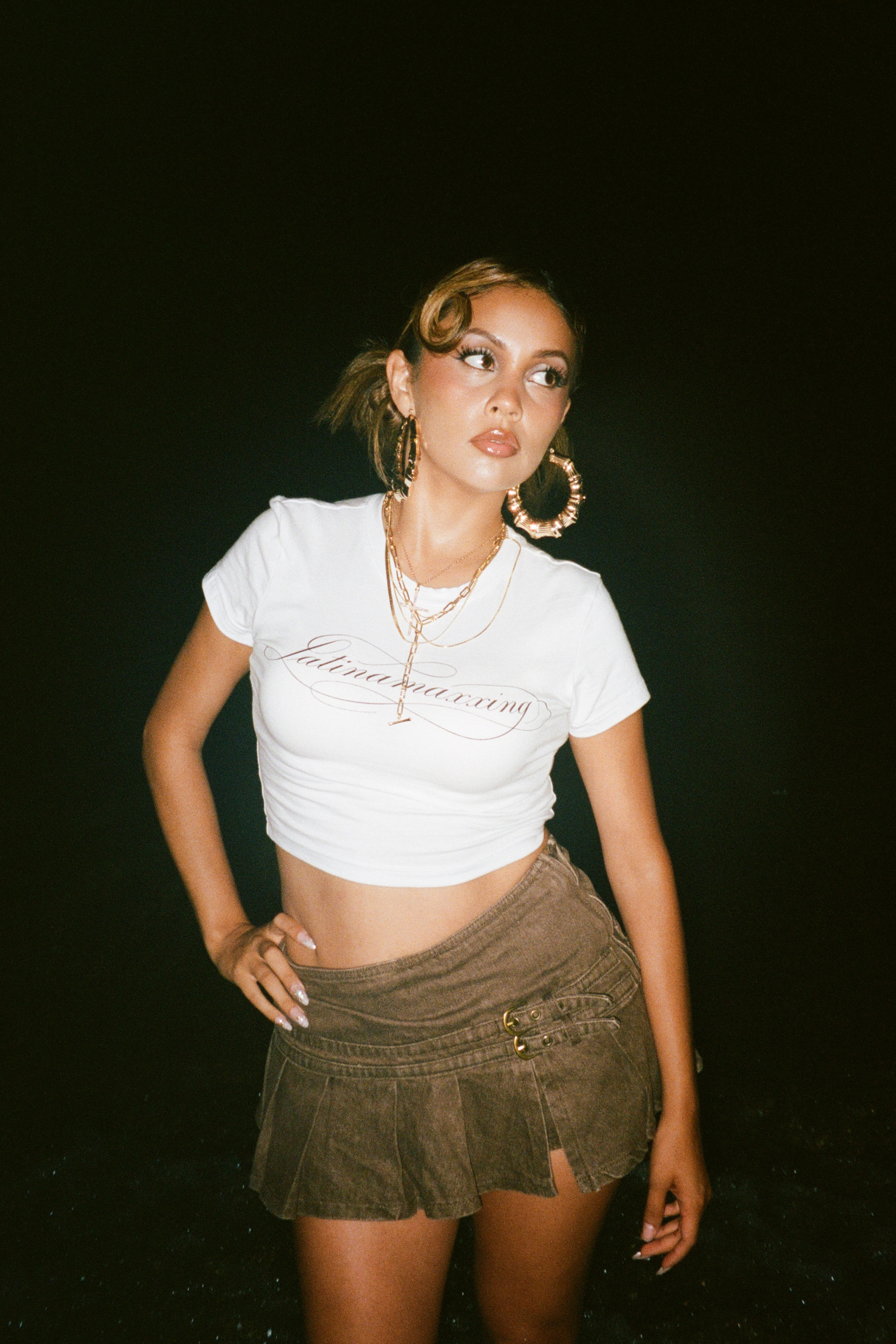 A woman posing in a white cropped graphic t-shirt and a brown corduroy mini skirt against a solid black background.